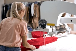Rear view on young caucasian blonde seamstress working on sewing machine in textile factory alone. Female controlling automated production of clothing in fabric. design, tailoring industry