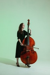 Girl musician with long hair in black dress plays the double bass against the light green background.