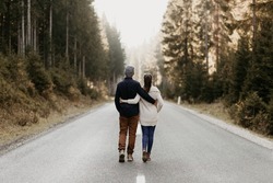 A couple in love is walking on an epmty road in the forest. Cuddling as they walk. Good relationship, active lifestyle. Together on the way. They face the distance. The background is bright and foggy.