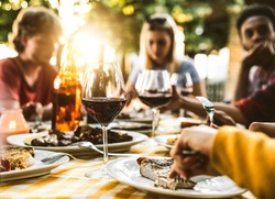 Group of friends having bbq dinner outdoor in garden restaurant - Multiracial family eating food at barbecue backyard home party - Focus on wine glass