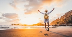 Happy man with hands up enjoying wellbeing and freedom at the beach - Male with backpack traveling in the nature with sunrise view - Healthy lifestyle, happiness and travel concept