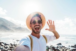 Young man with backpack taking selfie portrait outside - Smiling happy guy enjoying summer holidays at the beach - Millennial showing victory hands symbol to the camera - Youth and journey