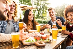 Group of multiracial friends having backyard dinner party together - Diverse young people sitting at bar table toasting beer glasses in brewery pub garden - Happy hour, lunch break and youth concept