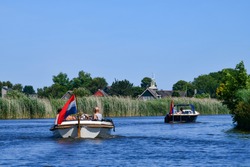 Low angle view from the water towards two recreational motor boats cruising along one of the canals lined with reeds beds between Frisian lakes in the Netherlands with a clear blue sky; 