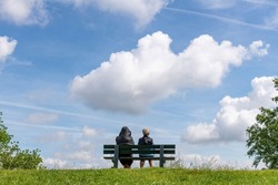 Low angle view of two people sitting on a park bench on top of a dike in the Netherlands looking in invisible distance with large cumulus clouds above in otherwise blue sky