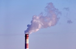 Smoking factory chimney. The plant's tall brick chimney emits a long cloud of thick white smoke against a blue sky.