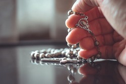 Close up of hand with the holy rosary to pray with light and shadows and reflection on the table in vintage tone. Beautiful Roman Catholic item, rosary made from silver. Life of faith concept.

