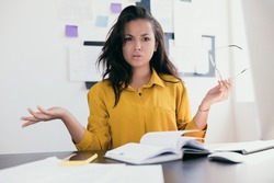 Perturbed office worker. Stressed young woman throw up her hands. Lady in yellow blouse holding glasses in hand. There is an open notebook and phone on the table. Office work work from home concept.