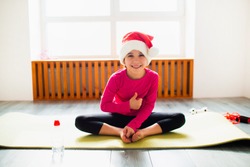Thumbs up Merry Christmas and a happy new year Little girl workout at home in santa christmas hat . Little girl workout at home. Little dark-haired female modelhas exercises near the window in room