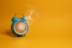 Hot coffee in a blue retro alarm clock on yellow background. Waking up with alarm and coffee concept.