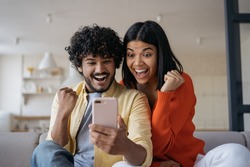 Excited friends using smartphone  playing mobile game relaxing at home. Happy emotional couple win online lottery celebration success. Multiracial students reading test results  