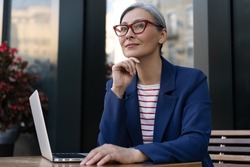 Pensive mature businesswoman using laptop computer working online at workplace. Portrait of middle aged grey haired manager wearing stylish eyeglasses planning startup. Successful business