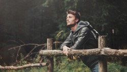 Young man with backpack hiking in the forest and leaning on a wooden fence, nature and physical exercise concept