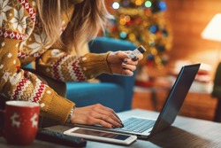 Woman doing online shopping at Christmas: she is sitting on the sofa at home, connecting with her laptop and holding a credit card, Christmas tree in the background