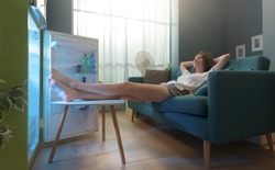 Happy woman lying on the sofa at home and cooling herself in front of the open fridge
