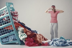 Disappointed angry woman with head in hands staring at a huge pile of dirty clothes falling from the laundry basket
