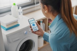 Woman using a smart washing machine, she is controlling the appliance from her smartphone