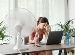 Woman sitting at desk at home during a summer heat wave, she is cooling herself with an electric fan
