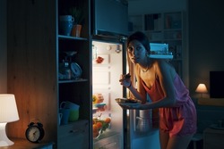 Hungry woman looking in the fridge and eating delicious pastries at night, she is breaking her diet