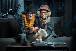 Frustrated couple sitting on the couch and feeling cold at home, they are trying to save money on their utility bills