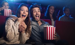 Young scared couple at the cinema watching an horror movie and screaming, she is holding her boyfriend's hand