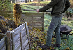 shuffling compost for aeration and better rot. the gardener uses a shovel and pitchfork in his hand. He has green pants and a jacket. Compost is in a wooden plank box. garden wheelbarrow