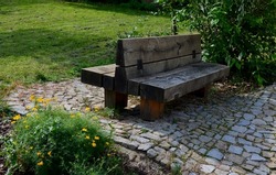 Park resting place with benches made of solid wood connected to a wooden log house pins. The benches are connected back to back. Lawn and flowering ornamental shrubs.