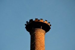 There is a metal toothed structure for the nesting of white storks on the brick historic chimney of the brewery. Brown burnt brick chimney. tin crown over the hole