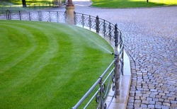 watering lawn automatic irrigation with pull-out sprinklers fresh green color black plastic nozzles extend and rotate in a circular rotation water granite cobblestone tiles and curb, fence, railings