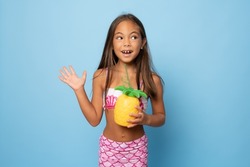 portrait of a little smiling girl in a swimsuit holding a pineapple on a blue background. Travel concept
