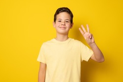 Smiling boy in casual t-shirt counting three with fingers isolated over yellow background.