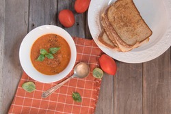 Tomato basil soup and grilled cheese sandwiches, fresh Roma tomatoes,  fresh basil, antique spoon and a folded  checked napkin on a wooden table.
