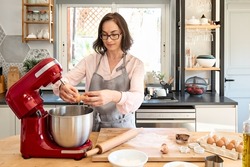 Woman wearing apron baking cookies in cozy kitchen. Housewife breaking eggs into a bowl of planetary mixer. Homemade pastries or cake.