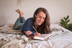 Woman in blue sweater lying on her bed writing in her leatherbound journal