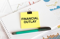 A yellow sticker with text Financial Outlay is in a Notepad with a green pen financial charts and documents. Business and financial concept
