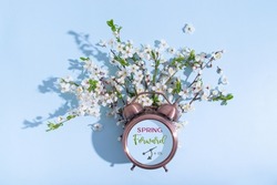 Spring forward text on alarm clock with instruction and blossom branches on a blue background. Flat lay, top view composition