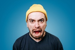 Close up young mad angry displeased guy in yellow cap looking at camera screaming. Young Caucasian angry man yells directly into the camera. Men emotions blue background, Furious man. Stress hysteria.