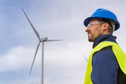 Renewable energy engineer looking at the sky with a wind power generator in the background. Maintenance operator with individual protection equipment in a wind farm. Leadership concept.