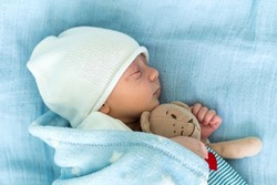 Close-up of Newborn Baby Face Portrait Early Days Sleeping With Tady Bear On Blue Background. Child At Start Minutes Of Life on Hat. Infants, Childbirth, First Moments Of Borning, Beginning Concept.