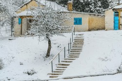 Traditional Cottage in a Snow Covered Village in a Greek Mountain During Snowfall on Winter Season. Beautiful Houses with Blue Windows and Doors. Stairs and Bare Tree in the Garden.
