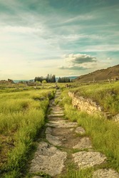 Beautiful retro vintage stone path. Stone ancient road with green tall grass on the side leading to the horizon under sunset golden sky. Abstract landscape of ancient city in nature. 