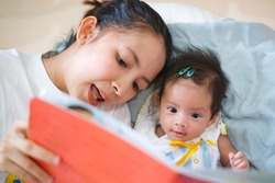 Smiling young Asian mother sit on the bed with little infant toddler child, happy biracial mom relax have fun read tale book with small baby girl at home, motherhood, childcare concept
