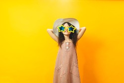 Beautiful kid girl in stripped dress, summer big hat and yellow star shaped big sunglasses posing with arms raised. Three quarter length studio shot on yellow background. Summer celebration concept.