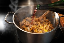 Red wine is poured from a bottle into a pot with roasting goulash to deglaze, cooking at home concept, selected focus, narrow depth of field