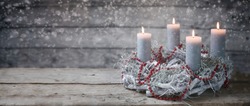 Advent wreath of white painted branches with burning candles and a red chain, time before Christmas, rustic wooden background with snowy bokeh and copy space, panoramic format, selective focus
