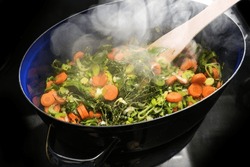 Steaming vegetables and herbs in a cooking pan on the black stove top, ingredients for a soup like carrots, celery, leek, thyme and bay leaves, copy space, selected focus, narrow depth of field