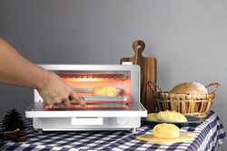 breads are put into white modern design toaster oven by right hand of a lady housewife , which is on the table with many baked homemade toast breads on grey cement wall background in the kitchen room