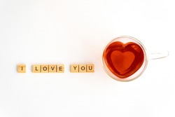 Heart-shaped glass teacup filled with red tea on a white background, wooden plates with the letters I LOVE YOU lined up to the left, horizontal, top view, free space
