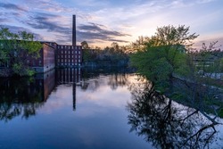 Sunset with reflection on the Nashua River. On the shore there is a historical building of a cotton factory with a tall brick chimney in an old industrial park. Nashua, New Hampshire, USA