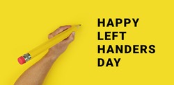 Big pencil in male left hand on yellow background with text Happy Left Handers Day. Conceptual banner for celebration of international left-handed day
